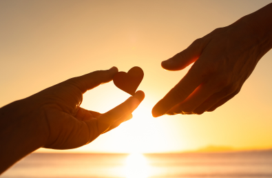 One hand holding a small heart and another reaching out with a sunset backdrop One hand holding a small heart and another reaching out with a sunset backdrop