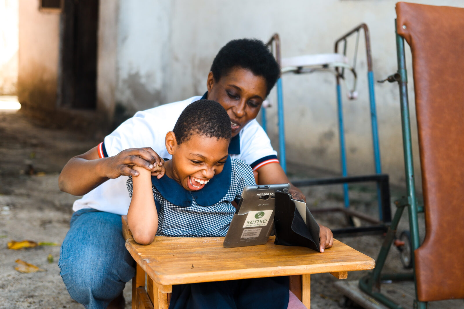Young black girl with deafblindness, smiling , sat down with a tablet on a table. He mum is behind her , holding her right hand. Young black girl with deafblindness, smiling , sat down with a tablet on a table. He mum is behind her , holding her right hand.