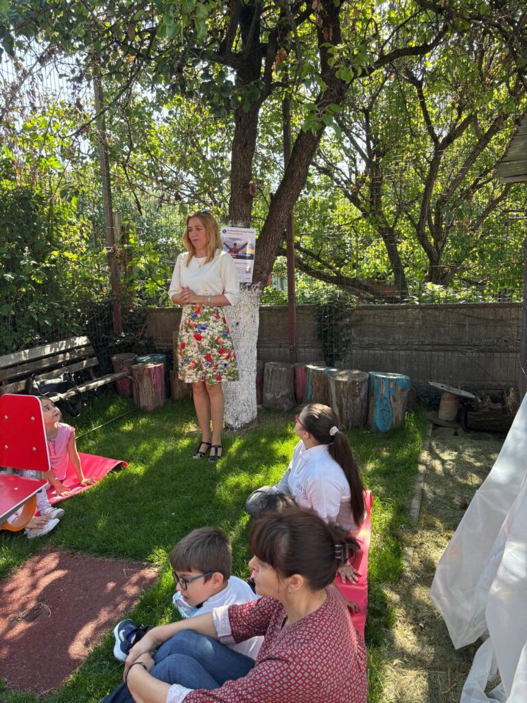 A woman stands, talking to a group of children and adults sitting on mattresses in a courtyard with trees. The atmosphere is calm, of educational activity carried out outdoors.