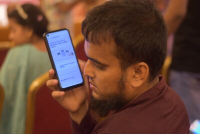 A young man with deafblindness, holding a mobile phone close to his ear as he listens to information from Insight. He is sat down in a conference room with other people n the background and he is wearing a maroon shirt. A young man with deafblindness, holding a mobile phone close to his ear as he listens to information from Insight. He is sat down in a conference room with other people n the background and he is wearing a maroon shirt.