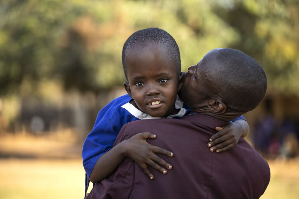 Pictured here is Karen with her father, Emmanuel. Emmanuel is carrying Keren and Keren is smiling facing the camera with her arms around her dad's shoulders. (Photo by Stuart Tibaweswa/Comic Relief)
