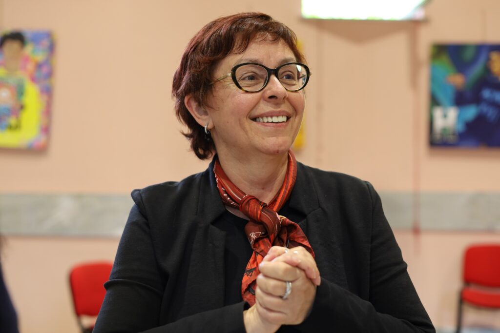 A close‑up photograph of a person standing indoors in what appears to be a community or conference space. The person is wearing a dark jacket over a top and a red patterned scarf tied around the neck. The person’s hands are held together at chest height, with fingers interlaced, and a ring is visible on one finger. The person’s face is intentionally blurred. In the background, red chairs are arranged along the wall, and colourful artwork or posters are visible, suggesting a meeting, event, or discussion setting. The image focuses on body posture and hand position, conveying active participation in a public or community environment.